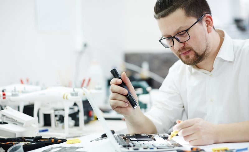 Side view portrait of man inspecting disassembled laptop with flashlight, looking for broken pats in workshop