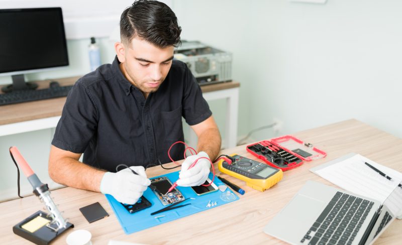 Hispanic male engineer checking the connections of a damaged smartphone with a multimeter at his repair shop
