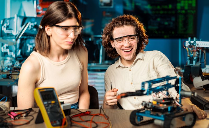 Excited young man and woman in protective glasses doing experiments in robotics in a laboratory. Robot and tools on the table